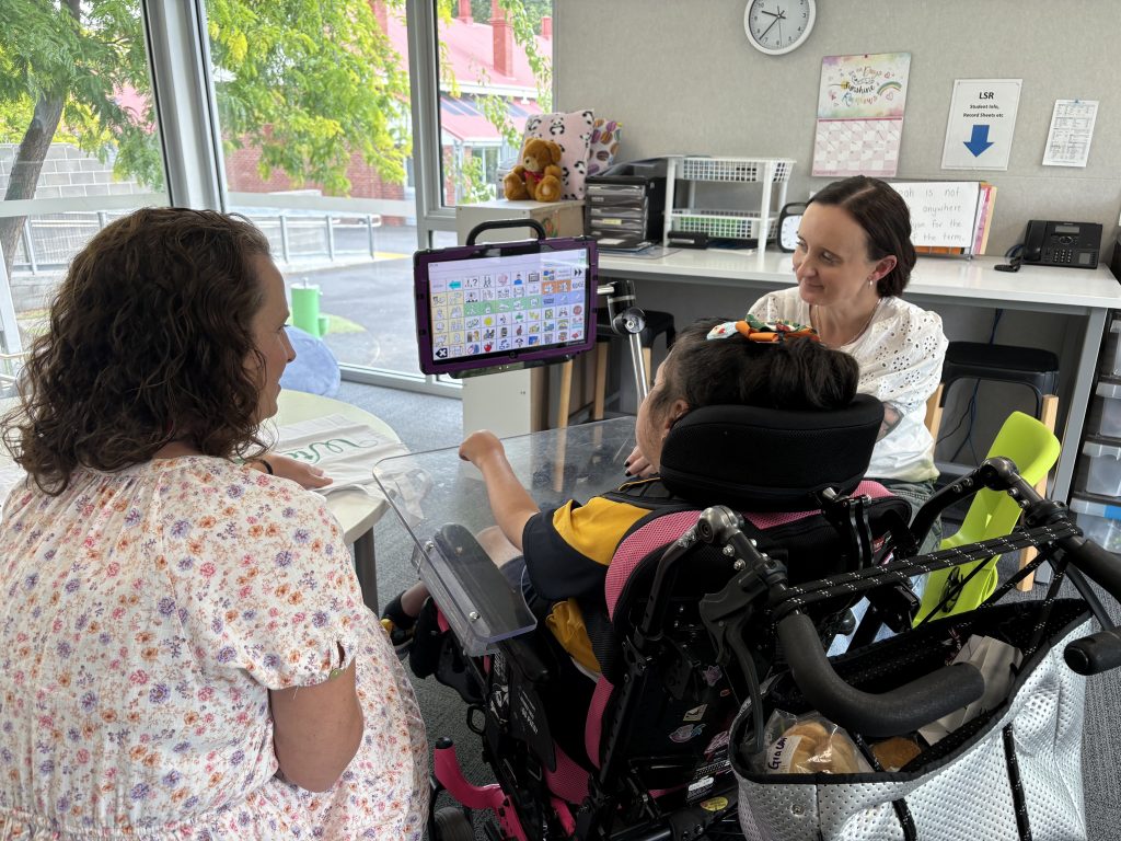 A girl in wheelchair using an Eyegaze to communicate. The Eyegaze has different symbols. 
Two women sit next to her. 